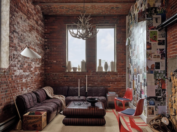 A cozy living room with a brick wall, a leather sofa, a wooden ottoman, a red chair, a floor lamp, and a wall of photographs.