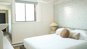 A bedroom with a large bed, a window, a nightstand with a lamp, and a mirror.