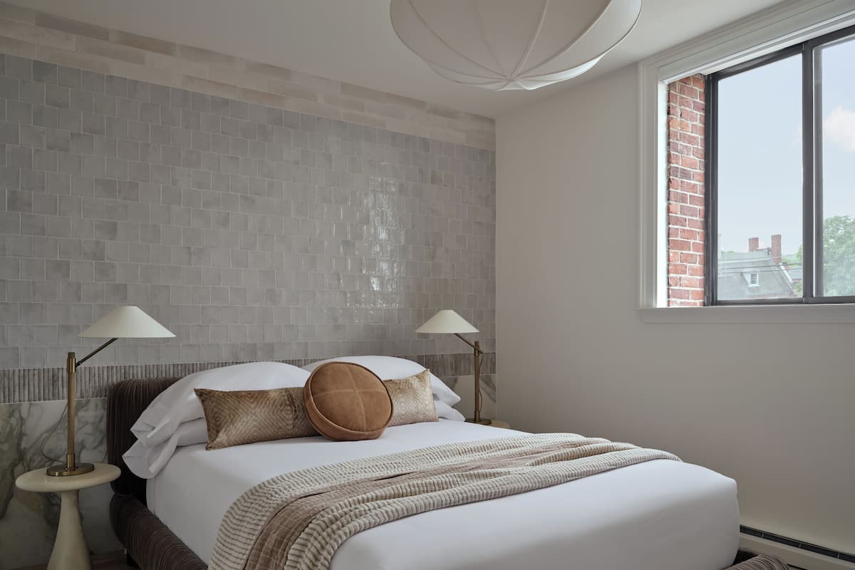A bedroom with a brick wall, a large window, a bed with white bedding, two bedside lamps, and a floor lamp.