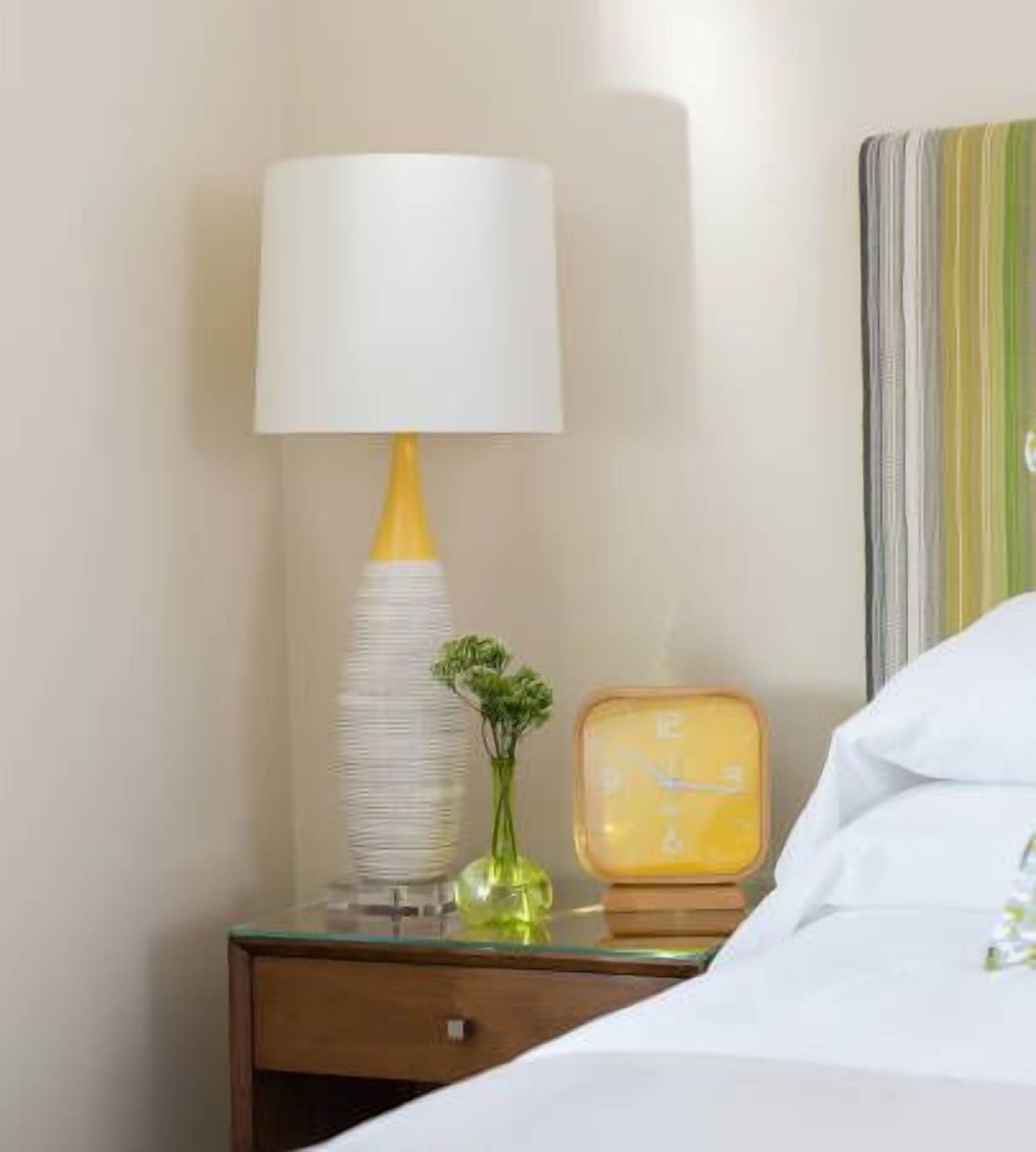 A neatly made bed with white linens, a bedside lamp with a white shade and yellow base, a small green plant, and a vintage-style alarm clock.