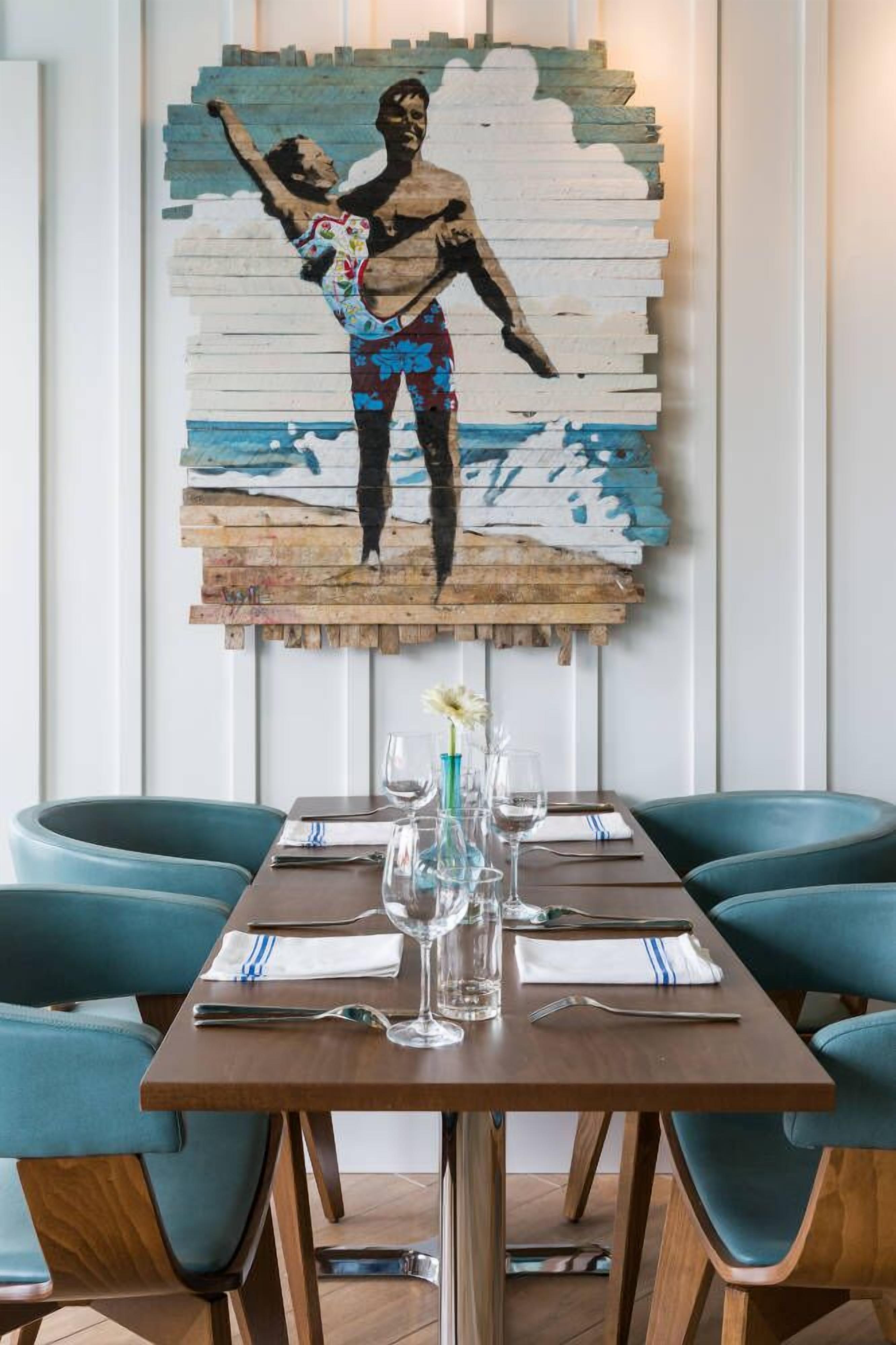 Ein Essbereich mit einem Holztisch, gedeckten Plätzen für zwei Personen, stuhlbezügen in Petrolfarbe und einem an der Wand hängenden Kunstwerk, das eine dynamische Figur zeigt.