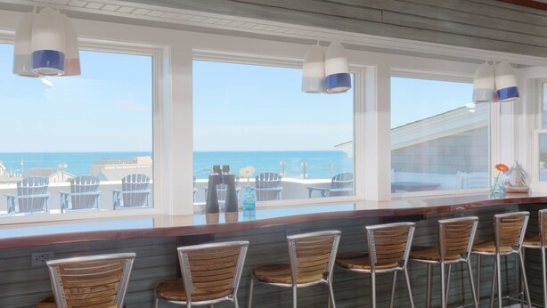 Un bar avec vue sur l’océan, des tabourets de bar et une fenêtre donnant sur les chaises longues et les parasols de la plage.