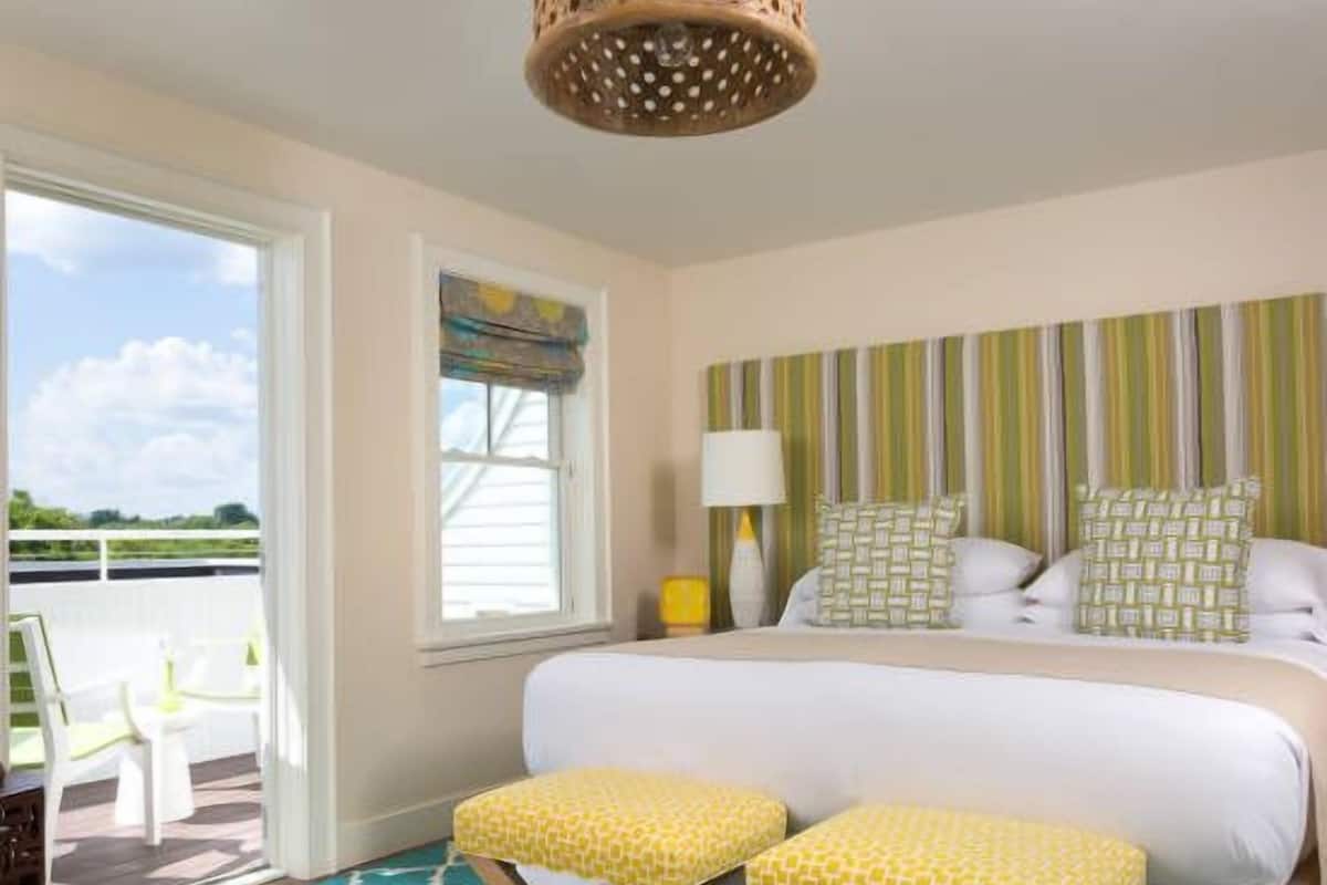 A hotel room with a large bed, two matching yellow ottomans, a window with a view, a striped headboard, and a hanging light fixture.