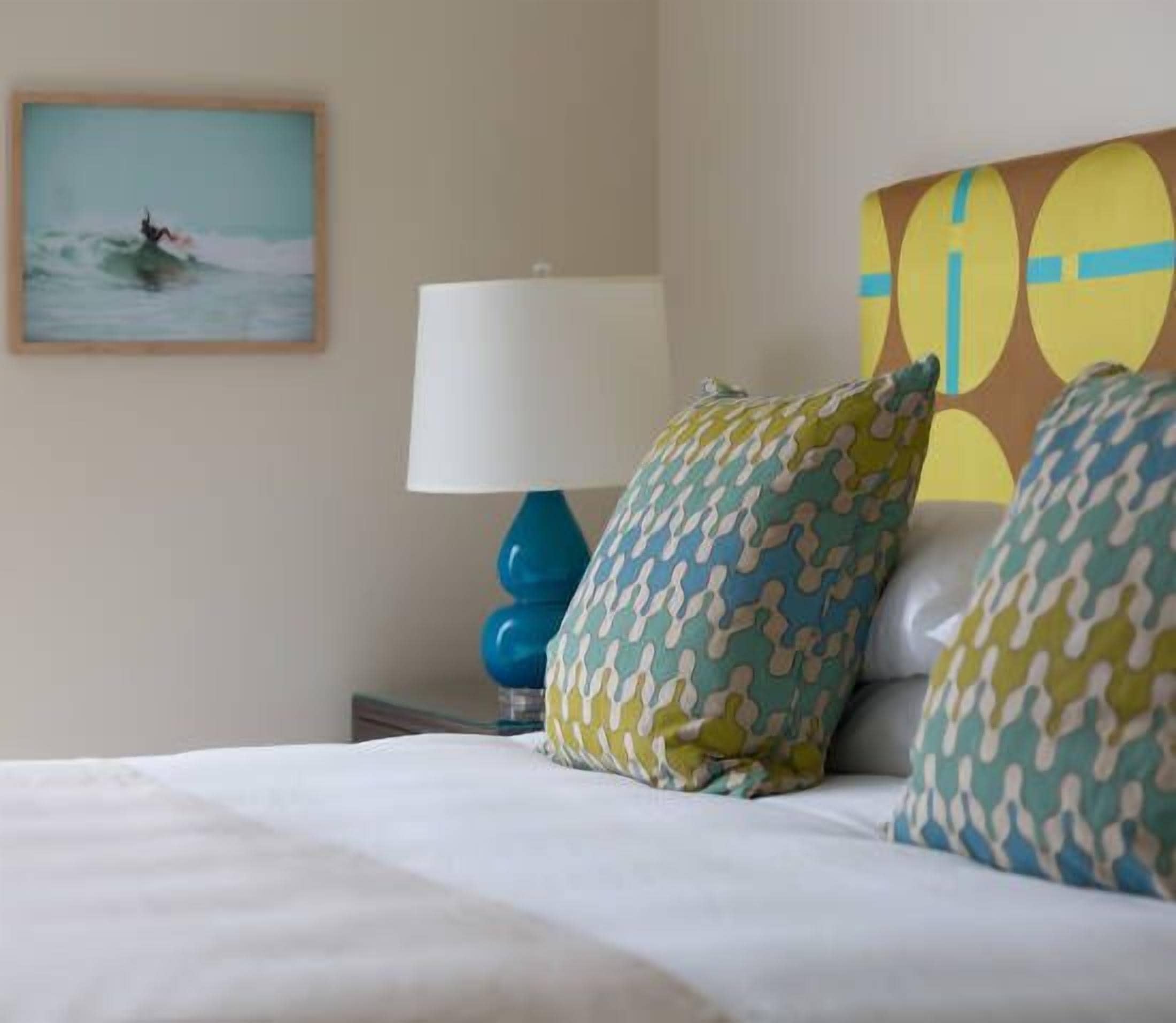 Une chambre d’hôtel avec un lit, une lampe bleue, des coussins à motifs, un tableau représentant un surfeur et une œuvre d’art abstraite et colorée accrochée au mur.