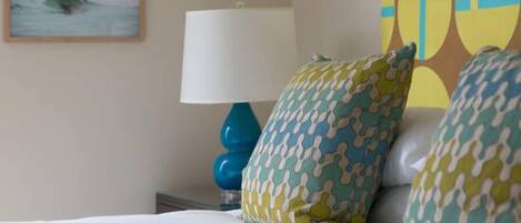 Une chambre d’hôtel avec un lit, une lampe bleue, des coussins à motifs, un tableau représentant un surfeur et une œuvre d’art abstraite et colorée accrochée au mur.