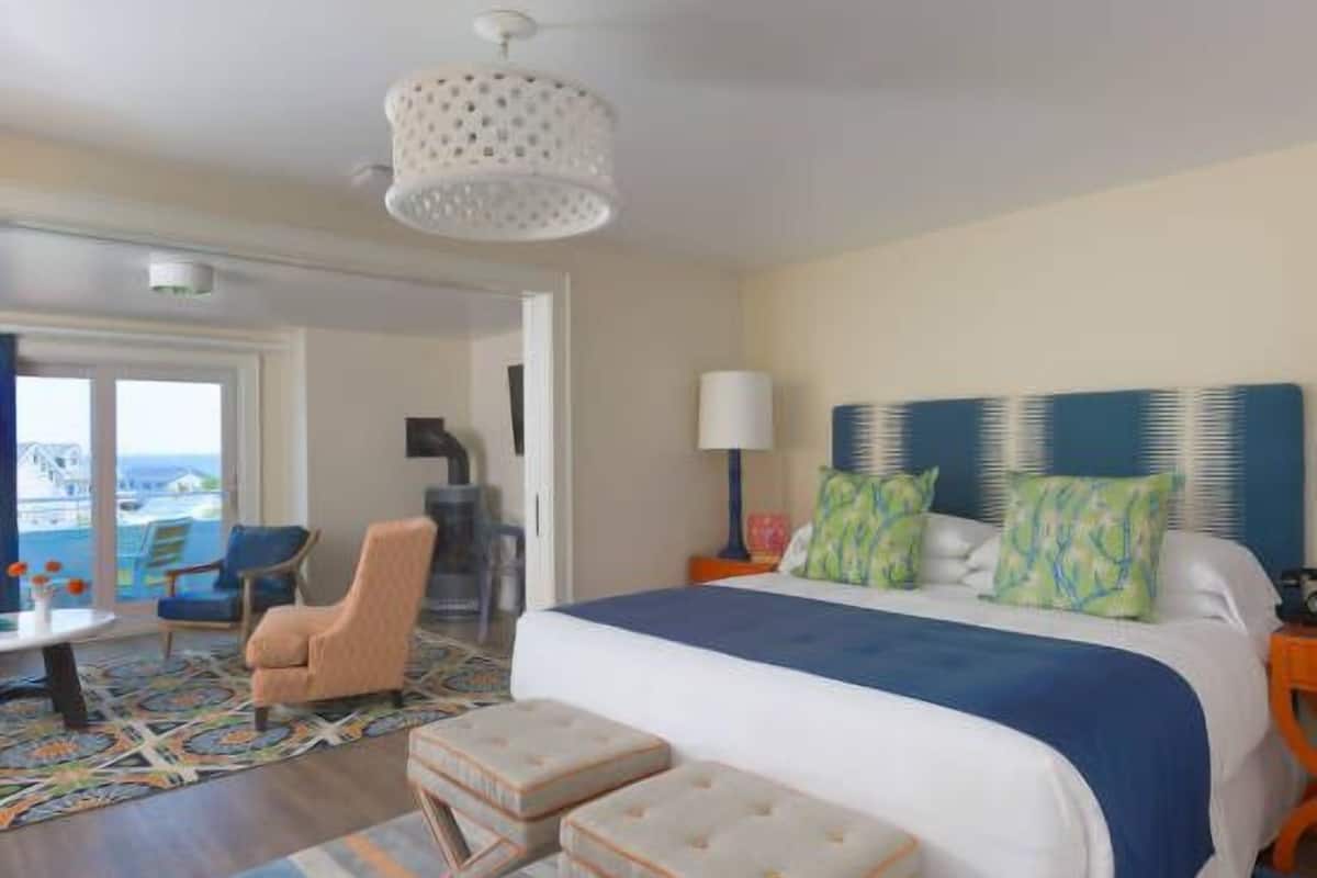 A hotel room with a large bed, two ottomans, a desk, a chair, and a view of the sea through the window.