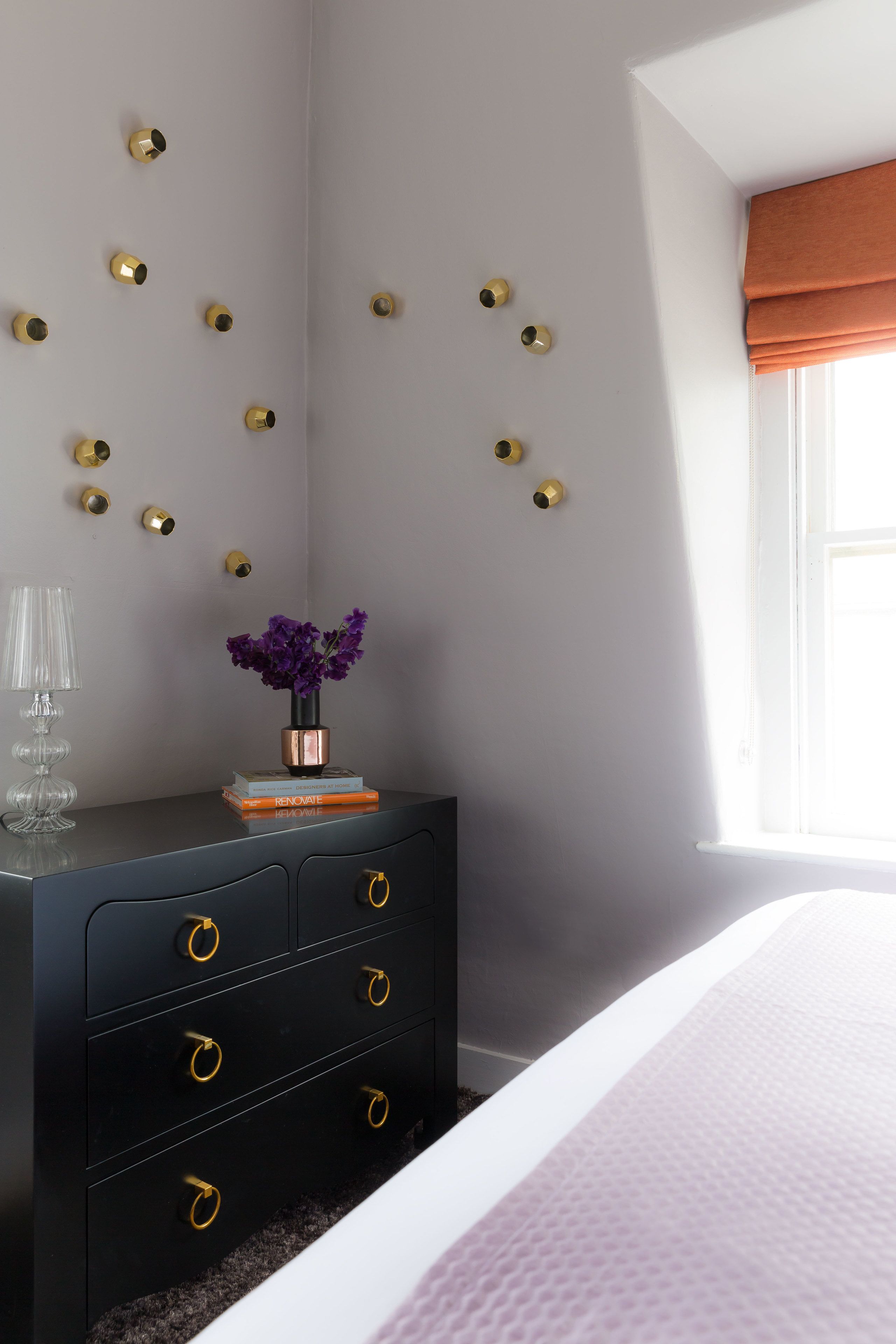 Een slaapkamer met een zwarte dressoir, een vaas met paarse bloemen en een raam met oranje gordijnen.