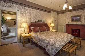 A bedroom with a large bed, two bedside lamps, a fireplace, and a door leading to another room.