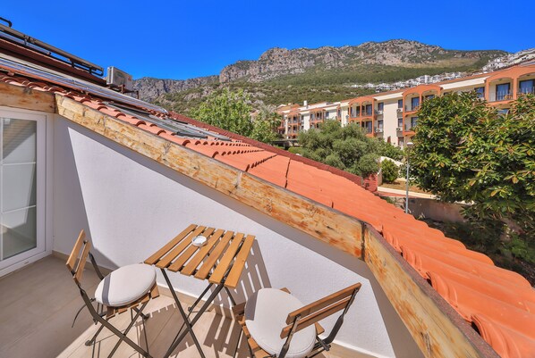 Un balcón con mesa y sillas de madera, con vista a un paisaje montañoso y edificios cercanos.