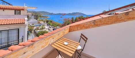 A balcony with a wooden table and chairs, overlooking a coastal view with buildings and a clear sky.