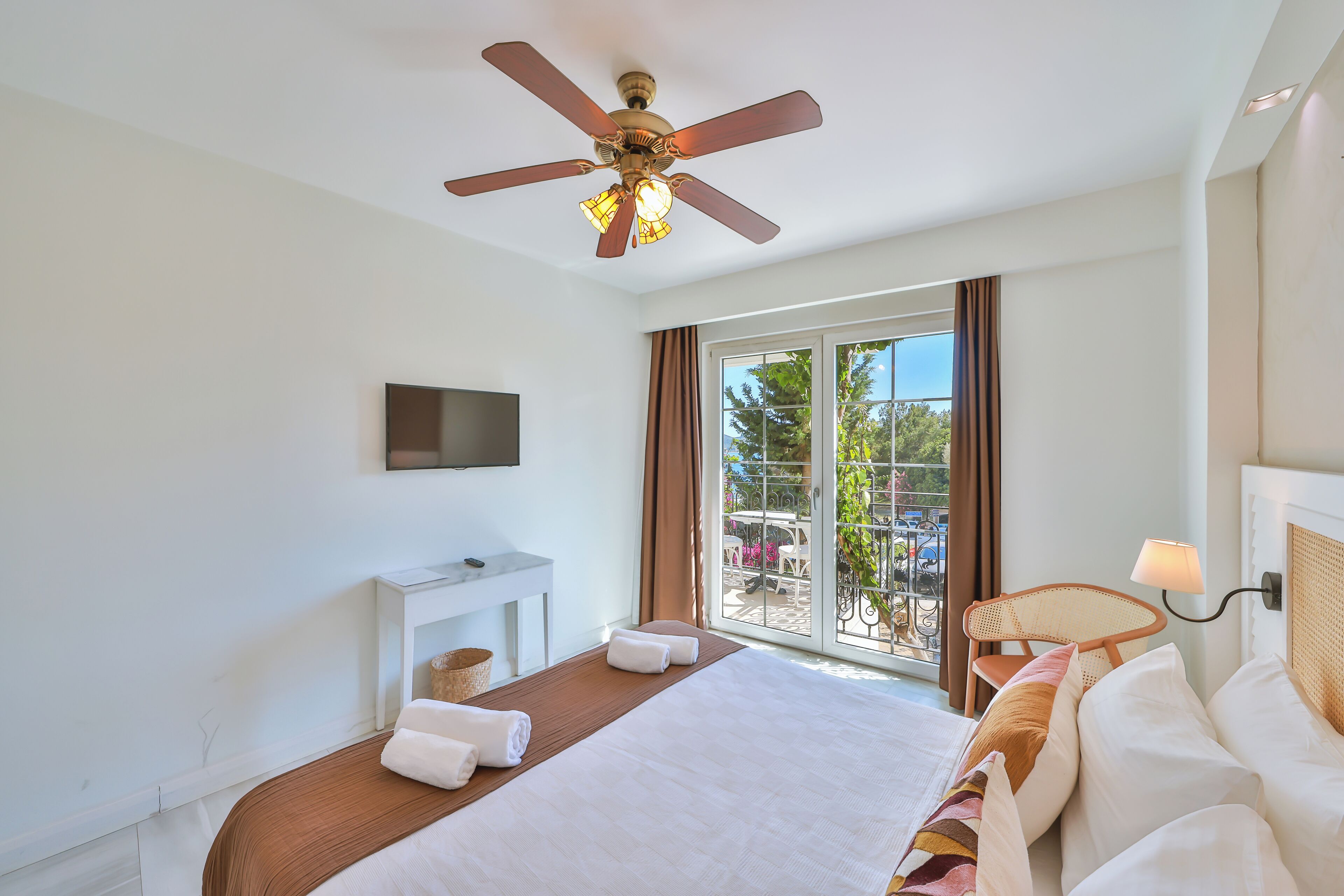Une chambre d’hôtel avec un lit, un ventilateur de plafond, une télévision, une fenêtre avec des rideaux, une petite table et une chaise.