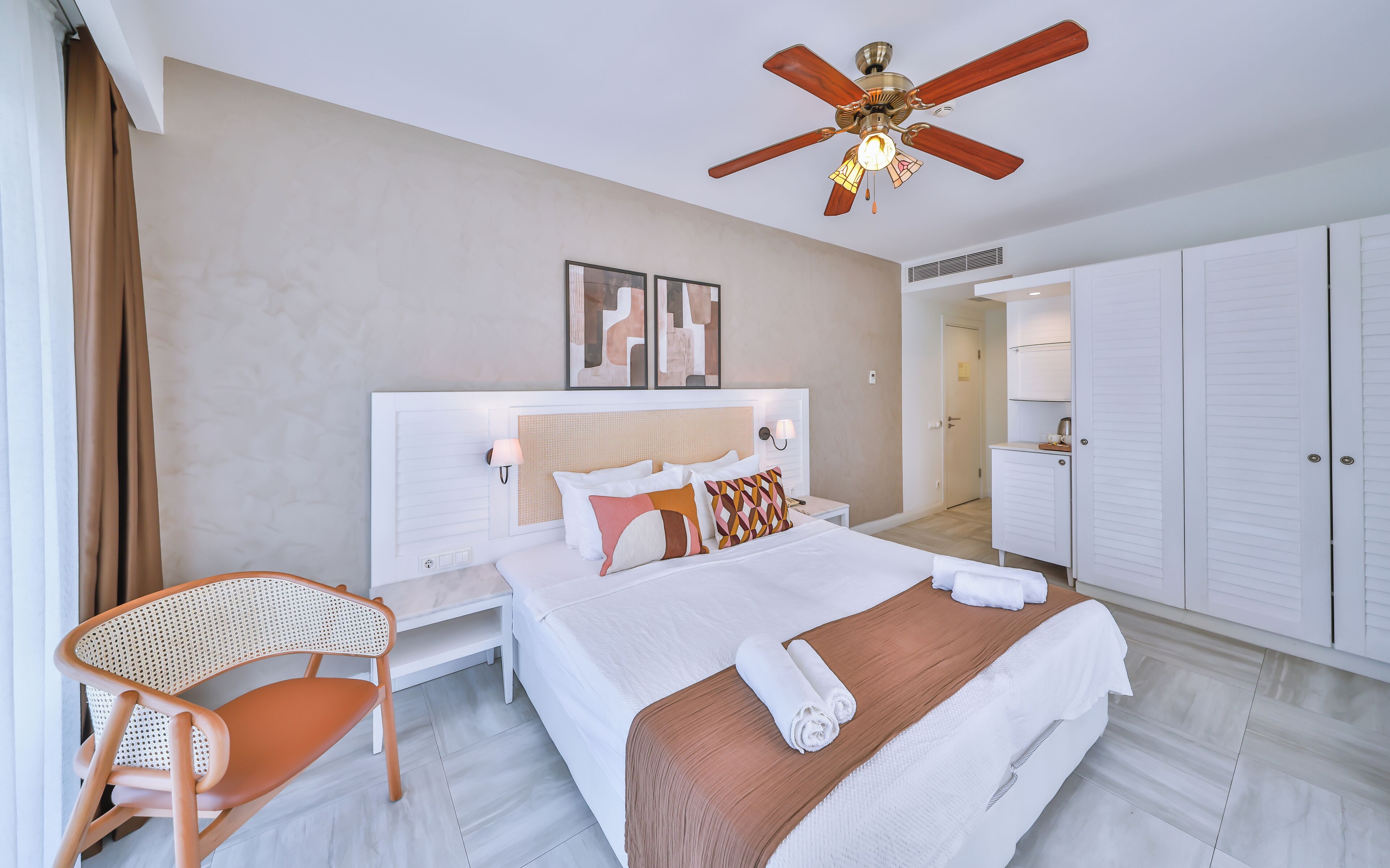 A hotel room with a bed, a chair, a desk, a wardrobe, and a ceiling fan.