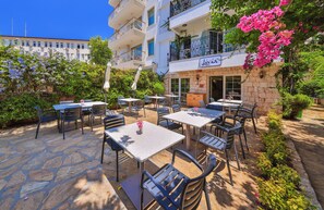 Un café al aire libre con suelo de piedra, sillas y mesas de metal, rodeado de vegetación y un edificio con balcón.