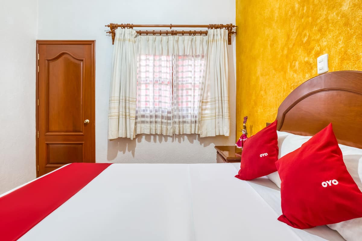 A hotel room with a wooden door, a bed with white bedding and red pillows, a window with curtains, and a yellow accent wall.