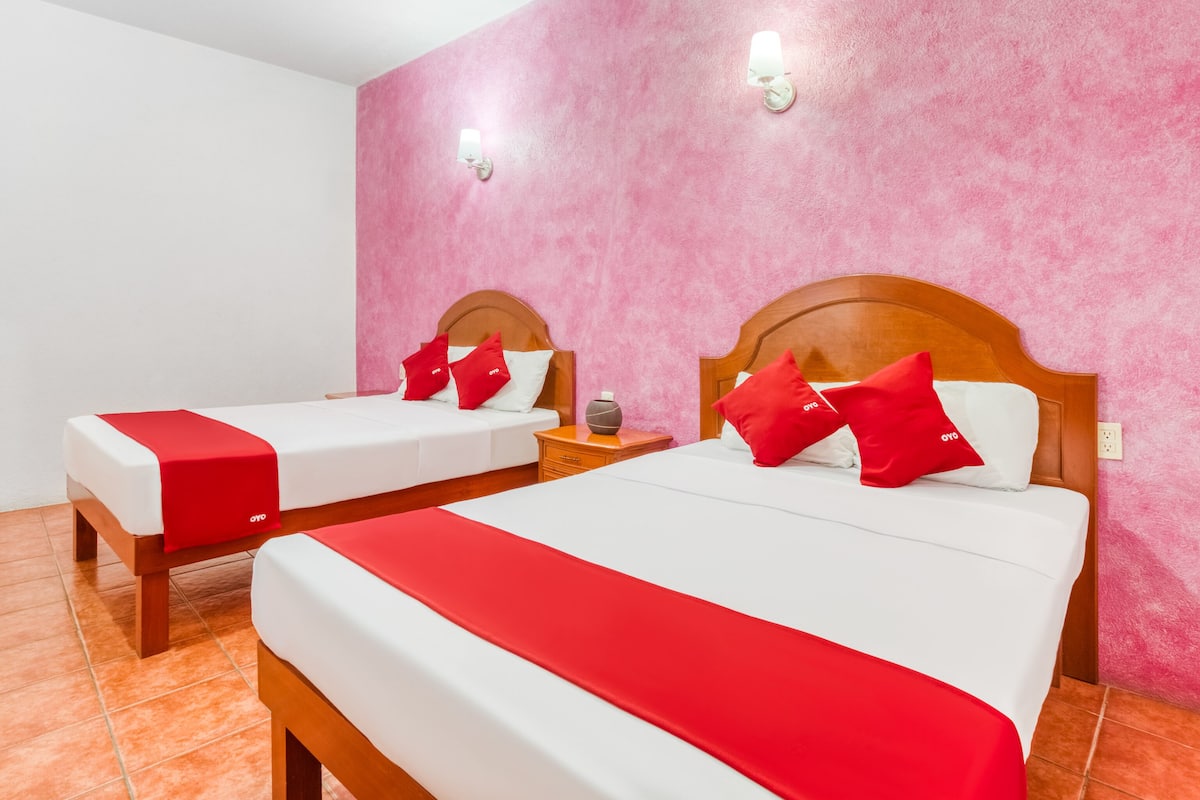 A hotel room with two beds, each with a wooden headboard, white bedding, and red accents. There are two wall-mounted lights and a nightstand with a lamp.