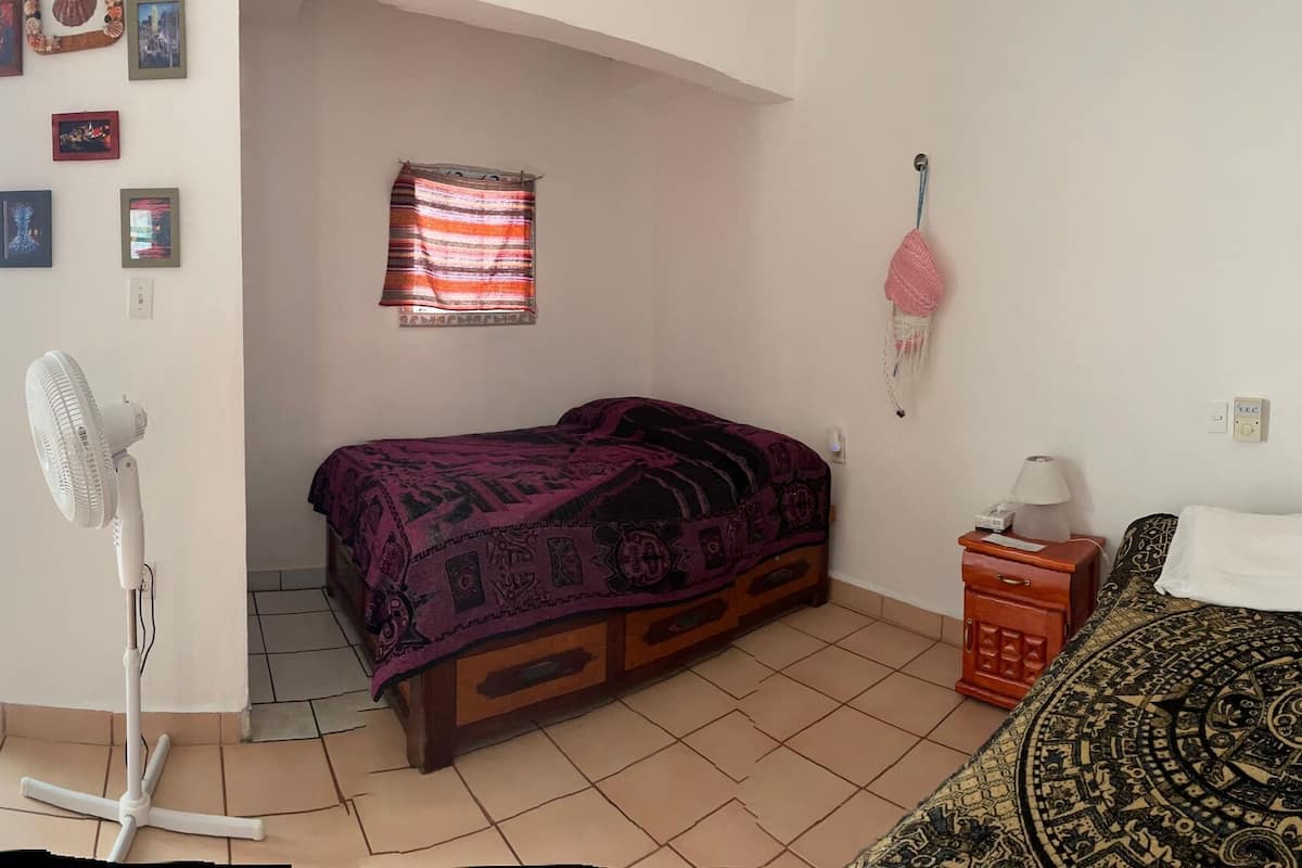 A small, well-lit bedroom with a bed, a fan, a nightstand, and a shelf with clothes.