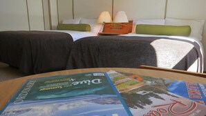 A hotel room with two beds, a wooden table with magazines, and a lamp on the nightstand. - Triple J Hotel & Cabins (Dawson City)