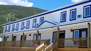A large, two-story building with a white facade and blue trim. It features multiple windows, a wooden balcony, and a staircase leading to the upper floor. - Triple J Hotel & Cabins (Dawson City)