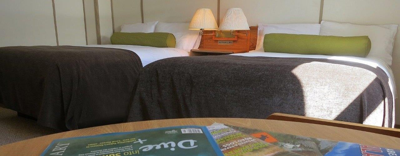 Una camera d'hotel con due letti, un comodino in legno e una rivista sul tavolo.