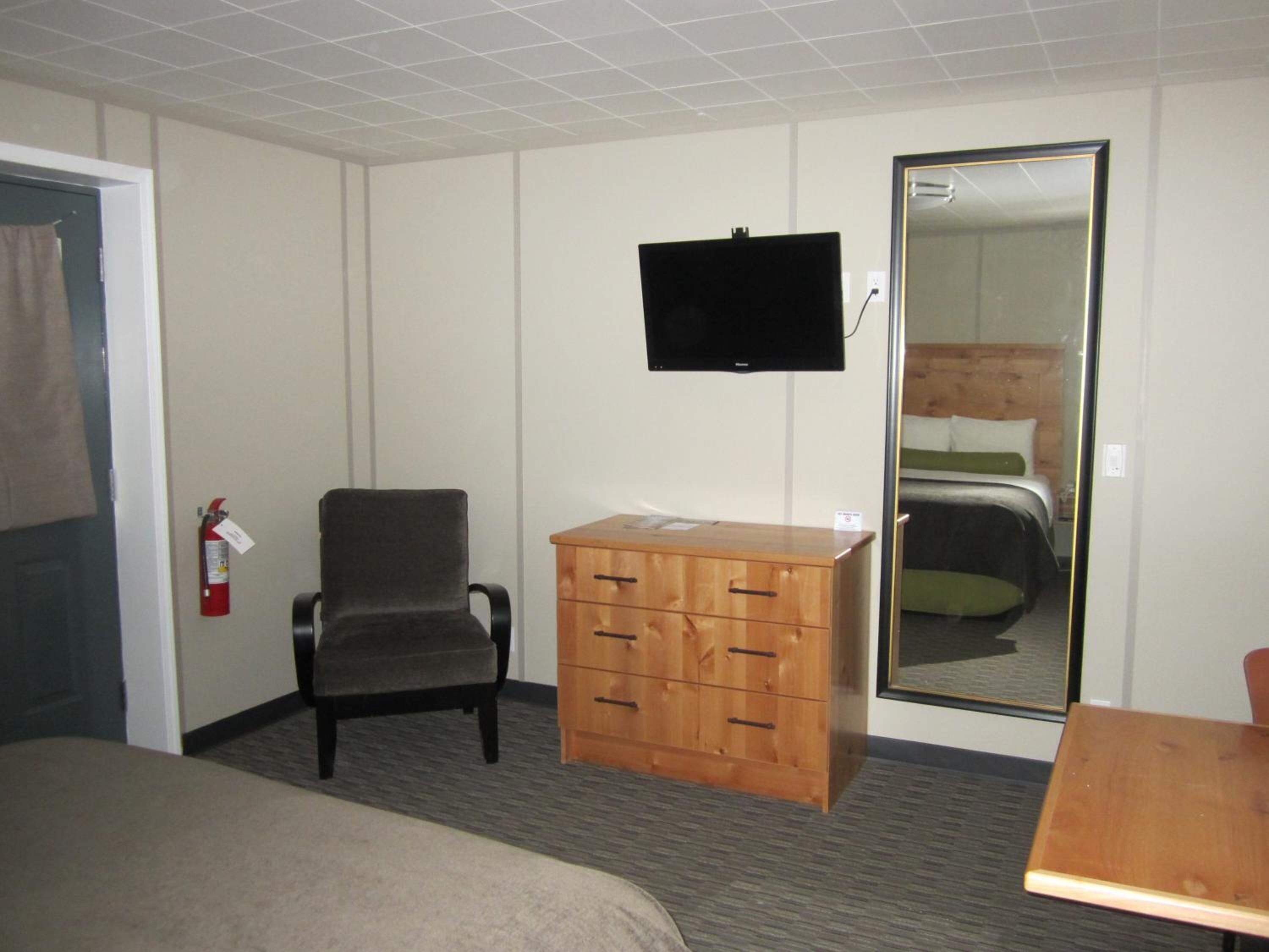 Una habitación de hotel con una cama, un aparador de madera, una silla, un televisor montado en la pared y un extintor.