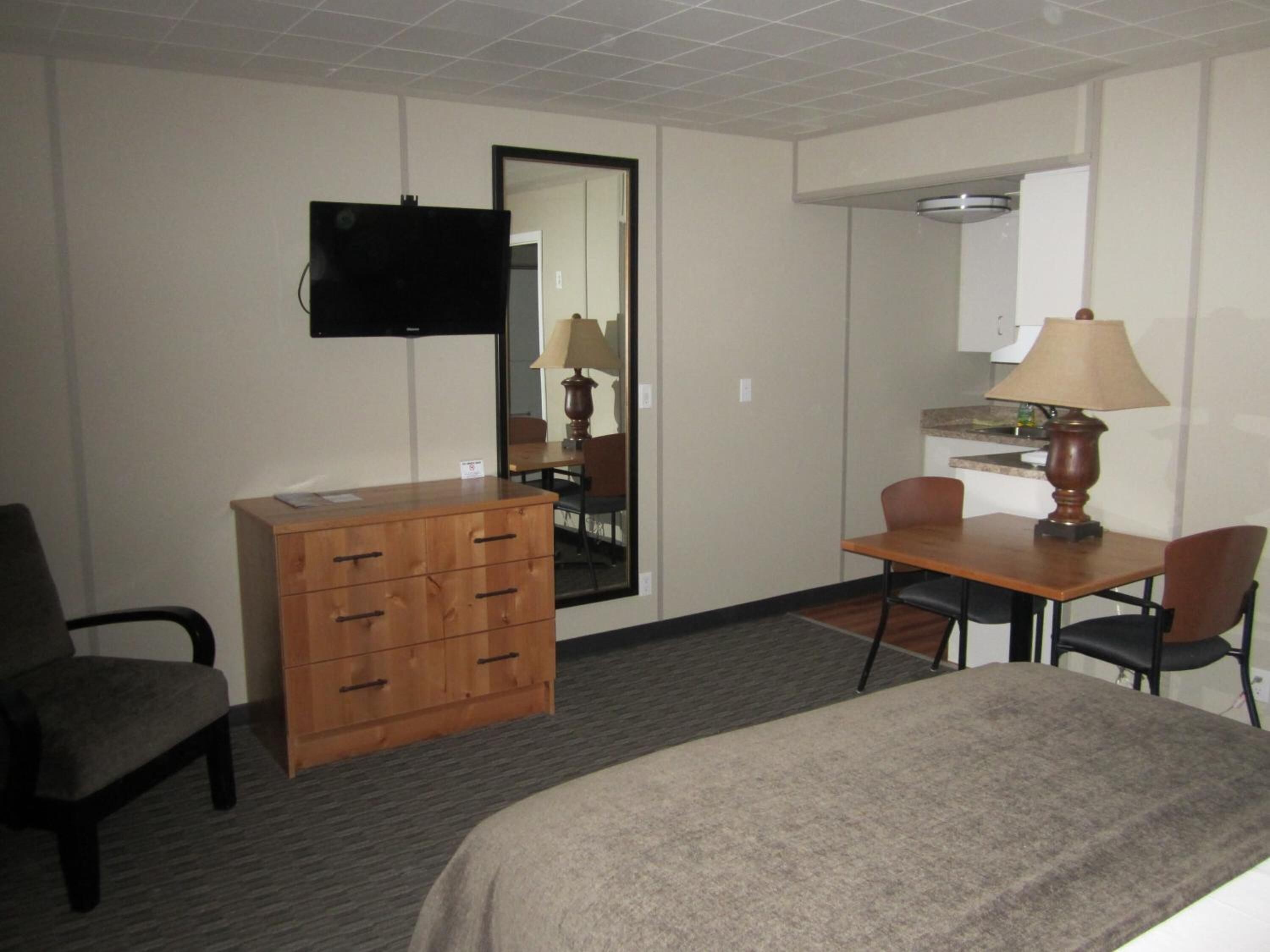 Quarto de hotel com cama, penteadeira de madeira, escrivaninha com luminária, cadeira e televisão na parede.