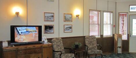 A hotel lobby with a wooden reception desk, two armchairs, a television displaying a room, framed pictures on the wall, and a sign with text.