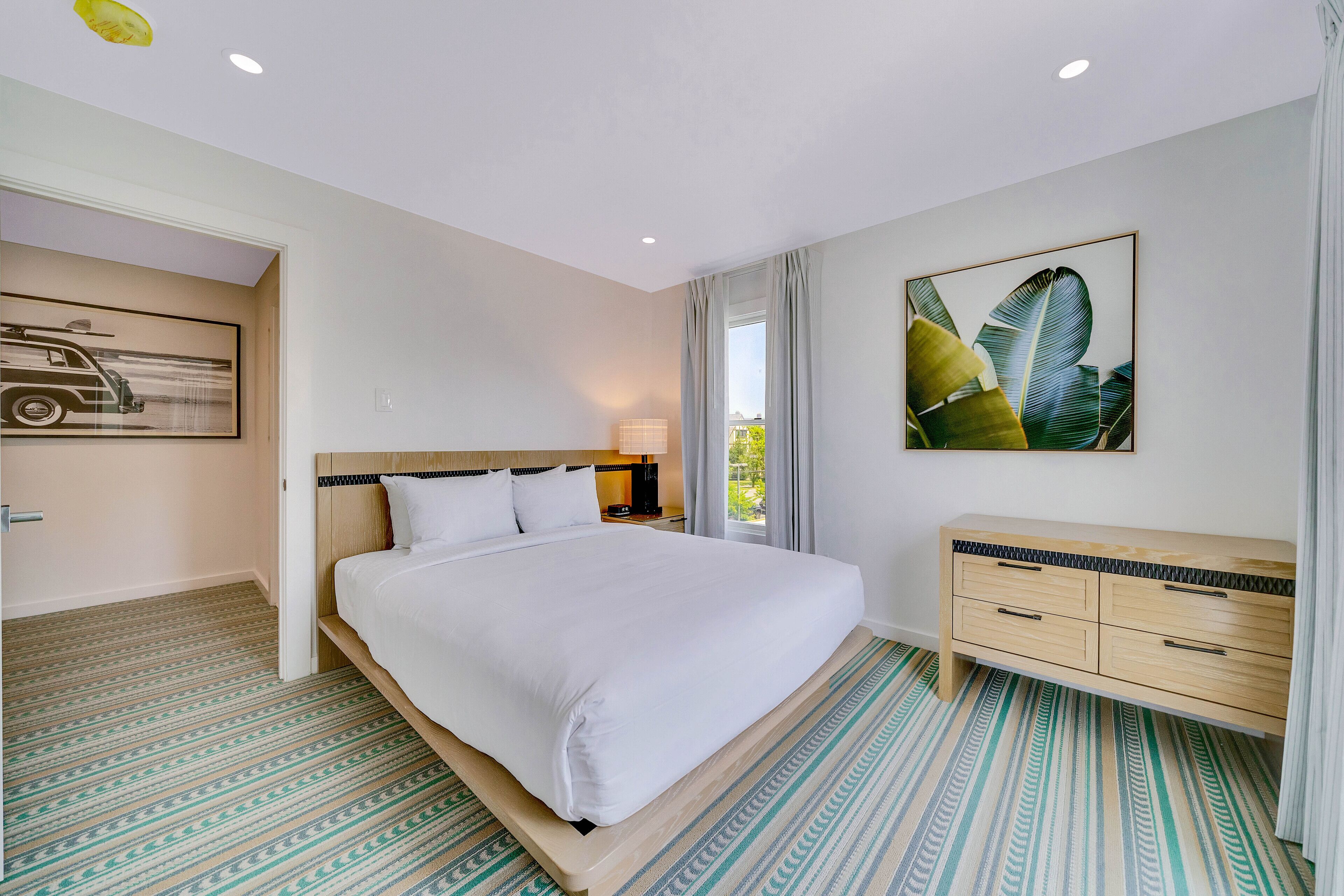 Une chambre d’hôtel avec un grand lit, une commode en bois, une table de chevet et un tableau représentant des feuilles.