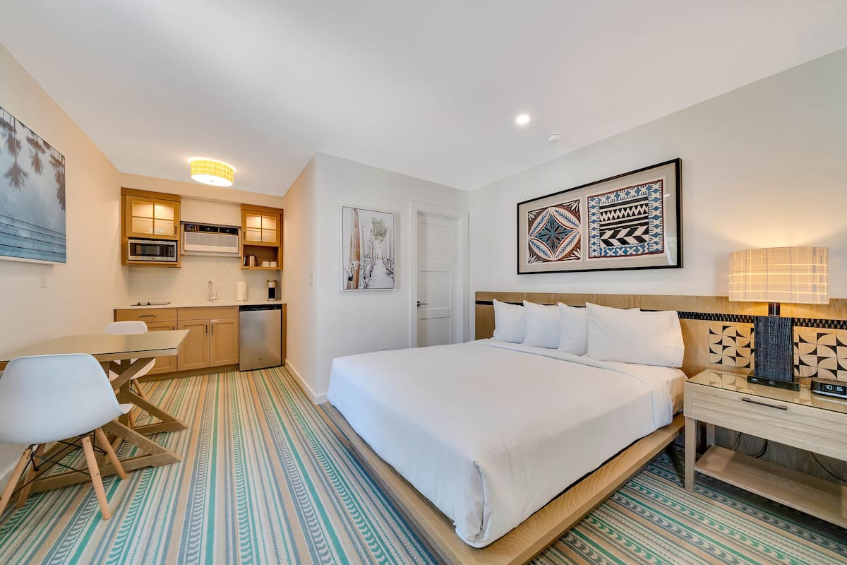 A hotel room with a bed, bedside table, chair, and a kitchenette area with a microwave and oven.