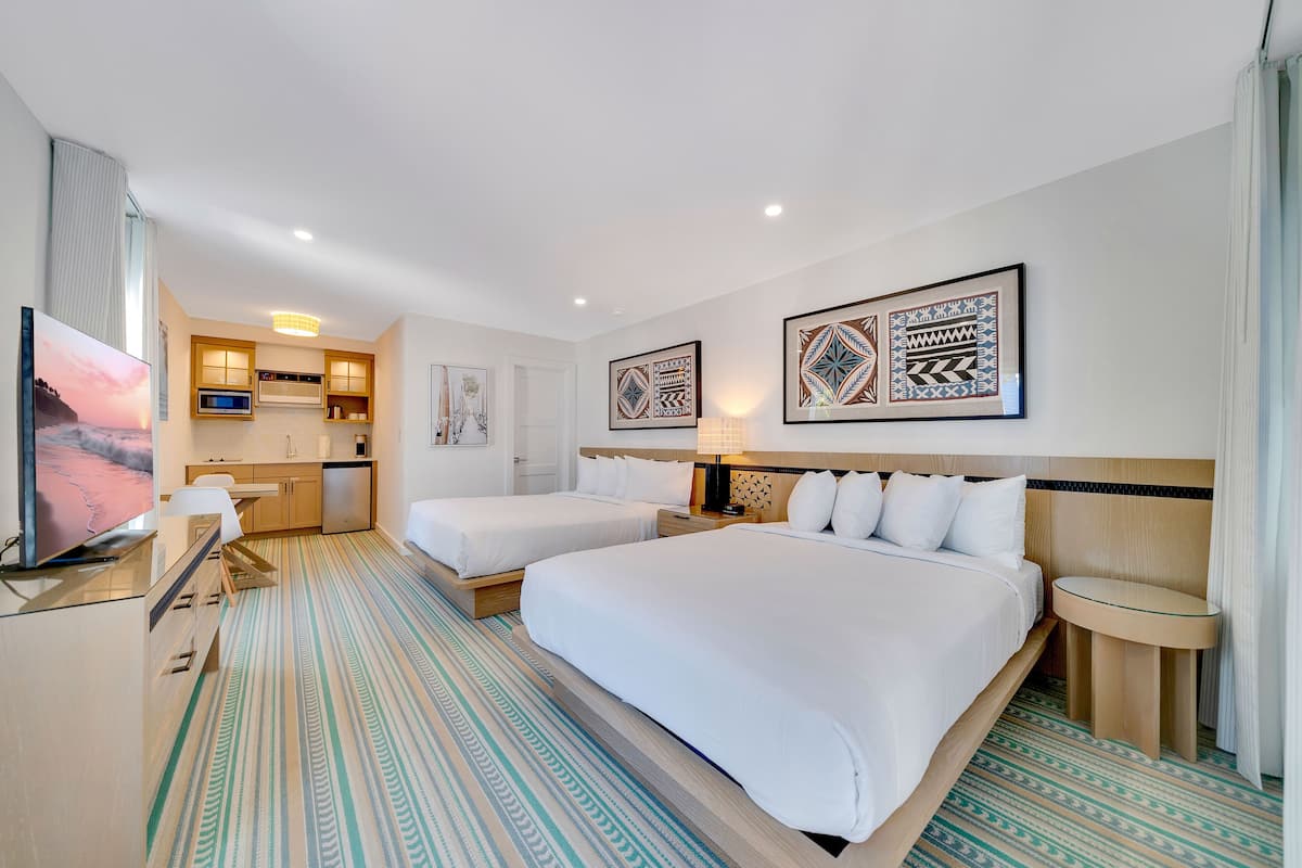 A hotel room with two beds, a desk with a TV, a kitchenette, and patterned artwork on the walls.