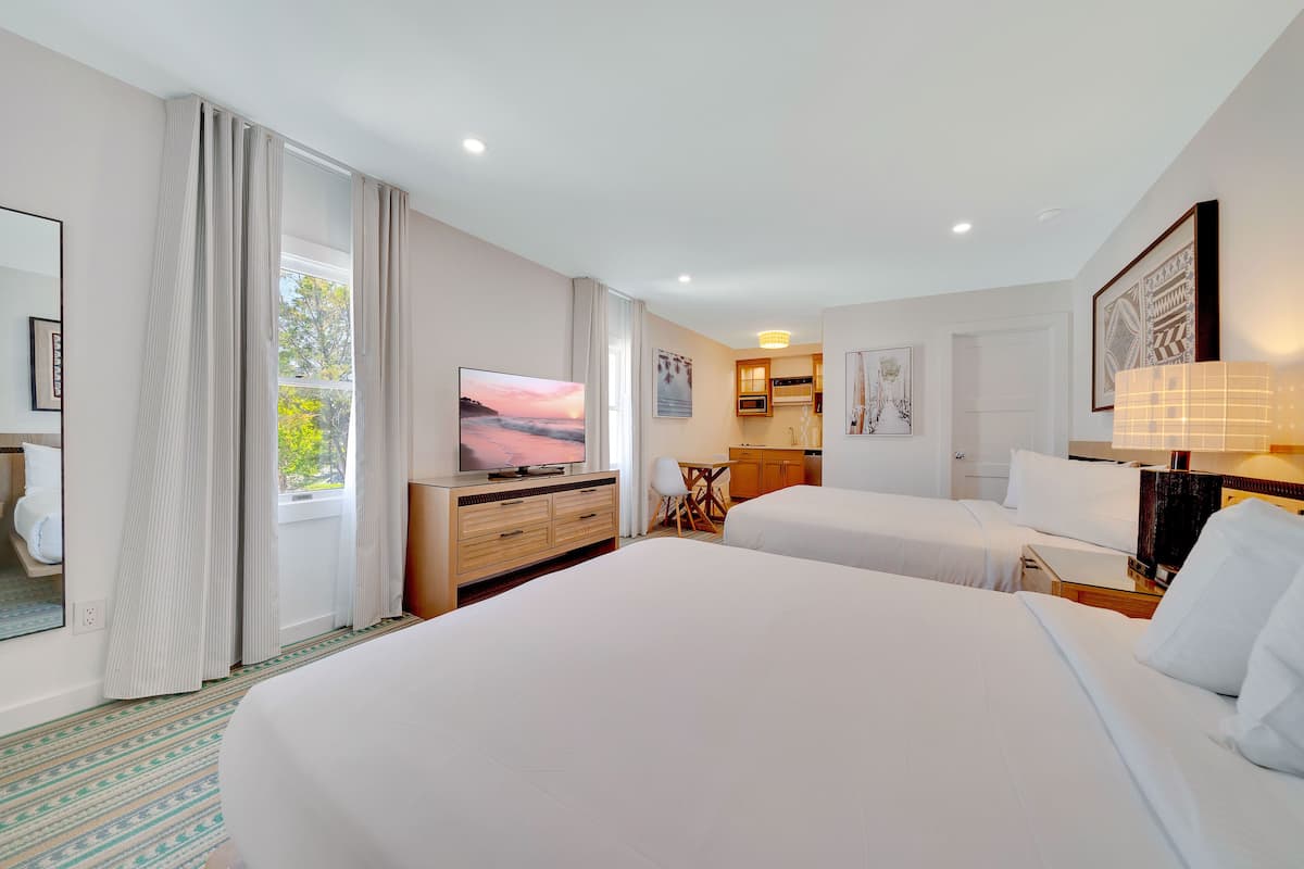 A hotel room with two beds, a TV, a desk, and a kitchenette.