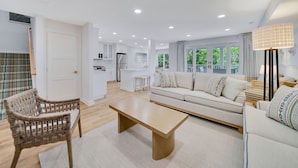 A modern living room with a sofa, a coffee table, and wicker chairs. There is a kitchen in the background with white cabinets and a dining area.