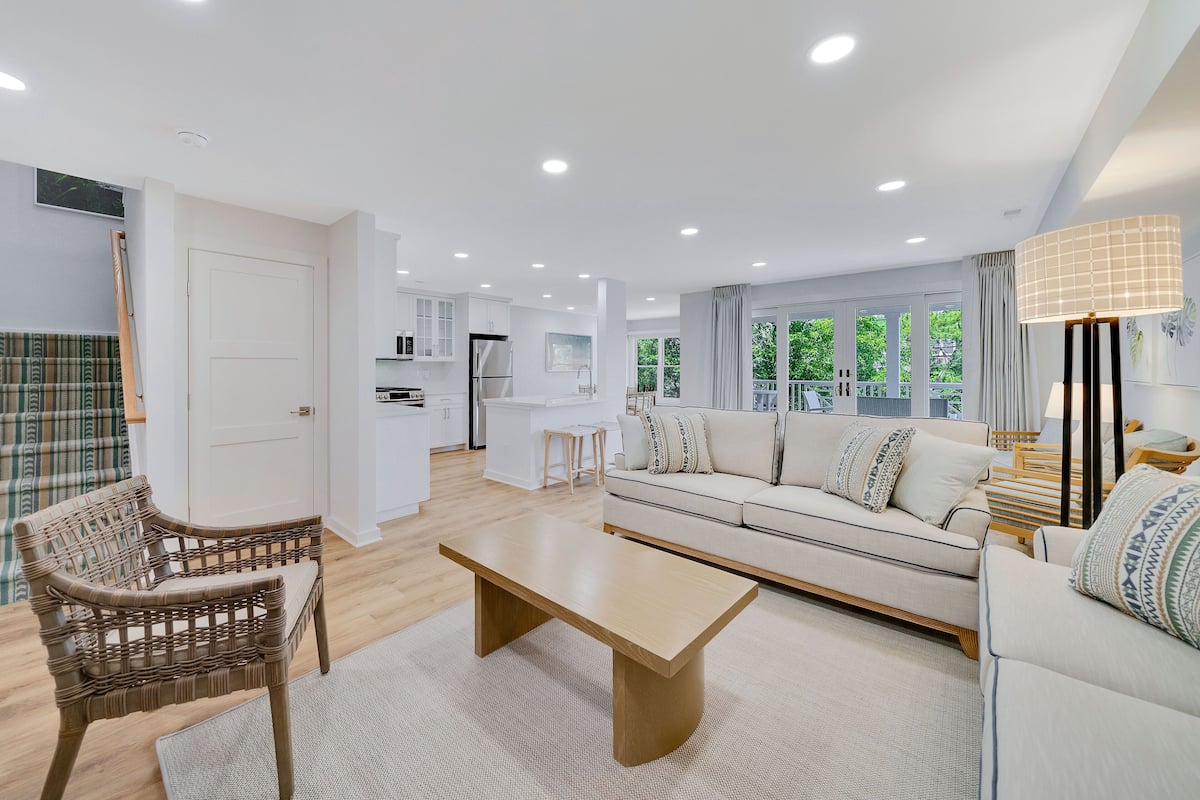 A modern living room with a sofa, a coffee table, and wicker chairs. There is a kitchen in the background with white cabinets and a dining area.