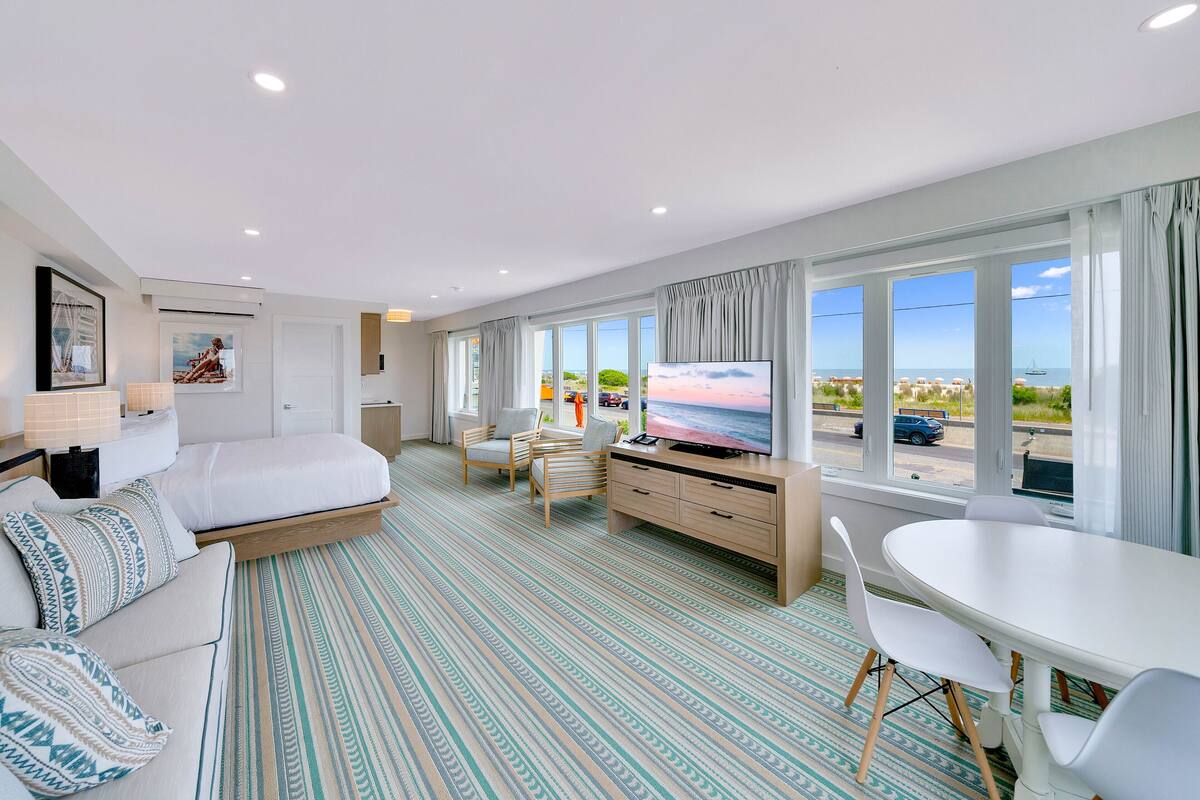 A modern hotel room with a bed, a desk, a TV, and large windows offering a view of the beach and city.