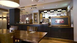 A restaurant with a wooden table and chairs, a bar area with a sign reading 'PROUD TO SERVE COSTA,' and a counter displaying various food items. - Premier Inn Aberdeen Airport - Dyce (Aberdeen)