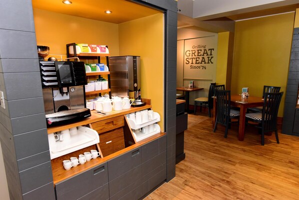 A coffee station with a coffee machine, cups, and a refrigerator. There is a dining area with a table and chairs. - Premier Inn Isle Of Wight - Newport (Newport)
