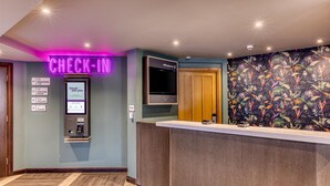 A hotel reception area with a neon 'CHECK-IN' sign, a digital kiosk, a television, and a floral-patterned wall. - Premier Inn Birmingham City - Aston (Birmingham)