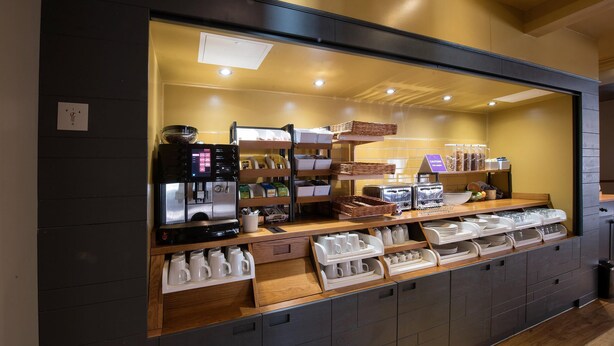 En bardisk i ett kafé med en kaffemaskin, hyllor med koppar och behållare samt ett utbud av bakverk.