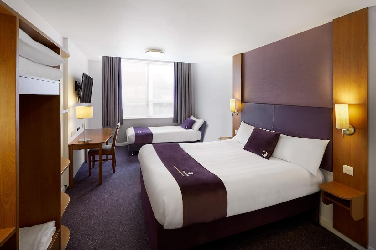 Ein Hotelzimmer mit einem großen Bett, einem kleineren Bett, einem Schreibtisch und einem Fernseher.