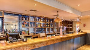 Een bar met een houten toonbank, flessenkasten vol drank en hangende pendelverlichting.