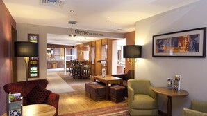A hotel lobby with a dining area, seating with lamps, a framed artwork, and a sign reading 'thyme eat. drink. relax.'