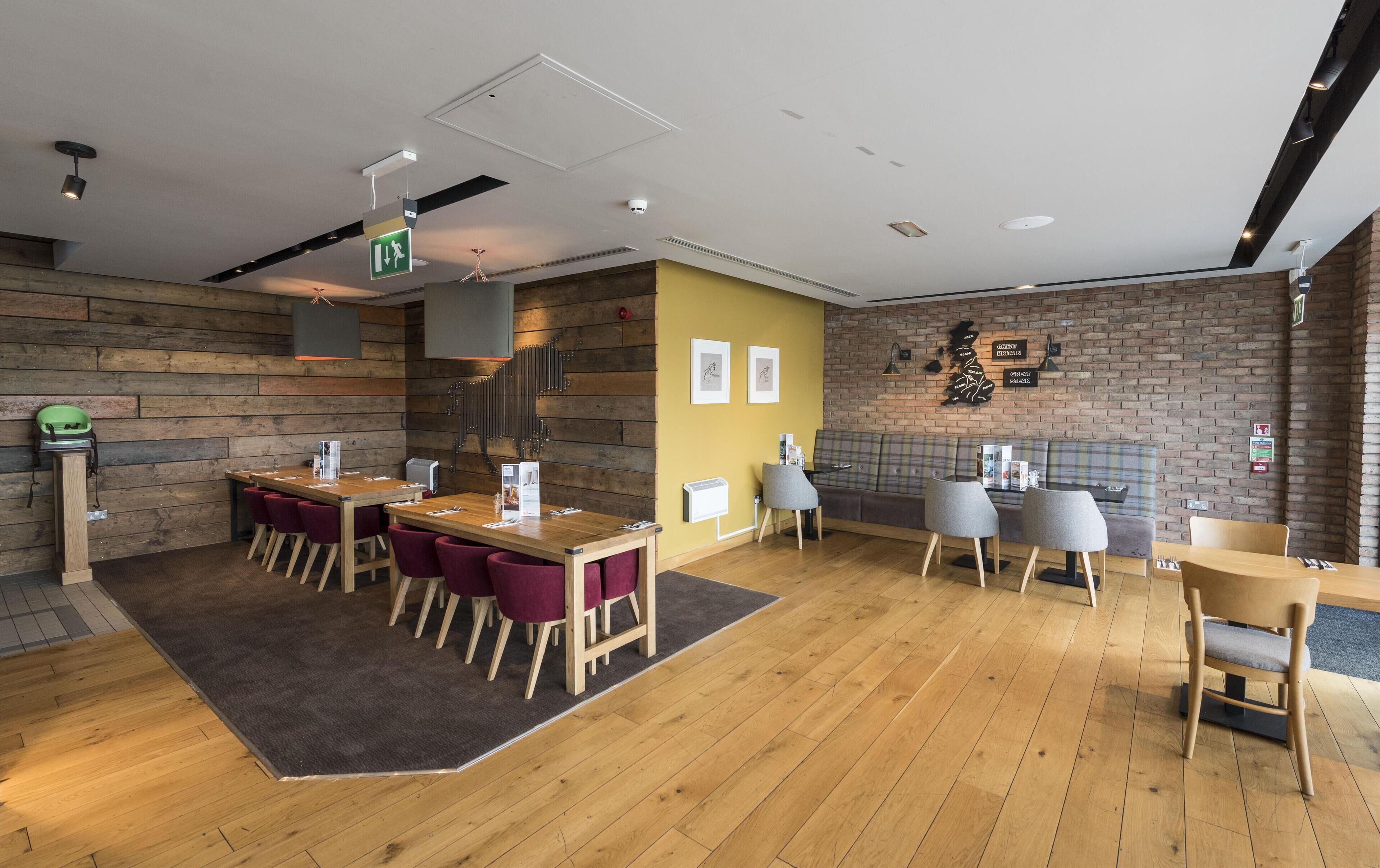 A modern restaurant interior with wooden flooring, a central dining table with chairs, a yellow accent wall, and a brick wall with artwork.
