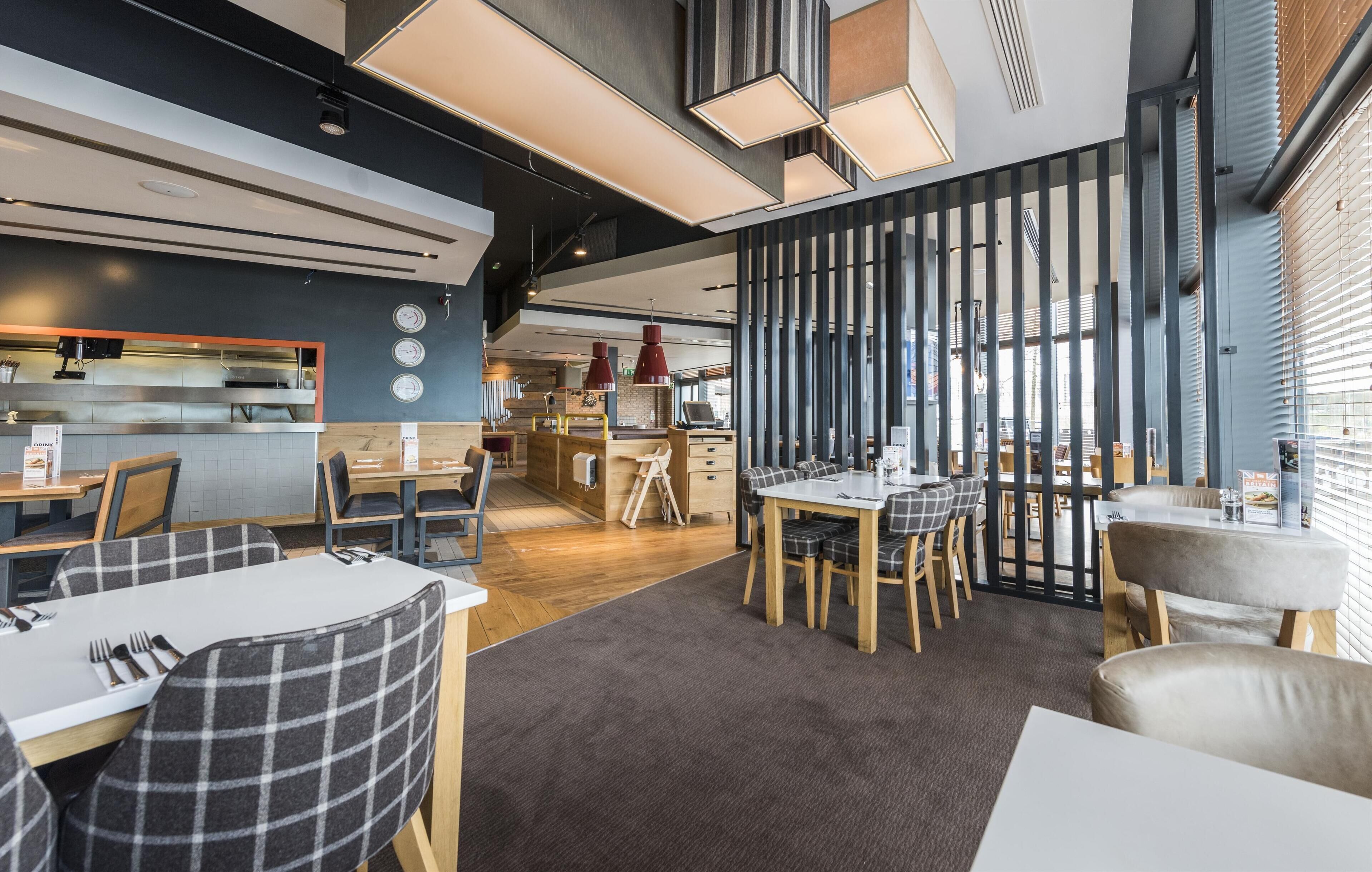 Ein modernes Restaurant mit einer zentralen Speisefläche, einer dahinterliegenden Küche und Blick auf die Stadt durch große Fenster.