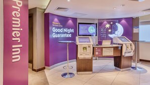 Zona de recepción de Premier Inn con pared morada, una pantalla digital mostrando un mensaje de bienvenida, un quiosco de autoservicio y un mostrador de recepción.