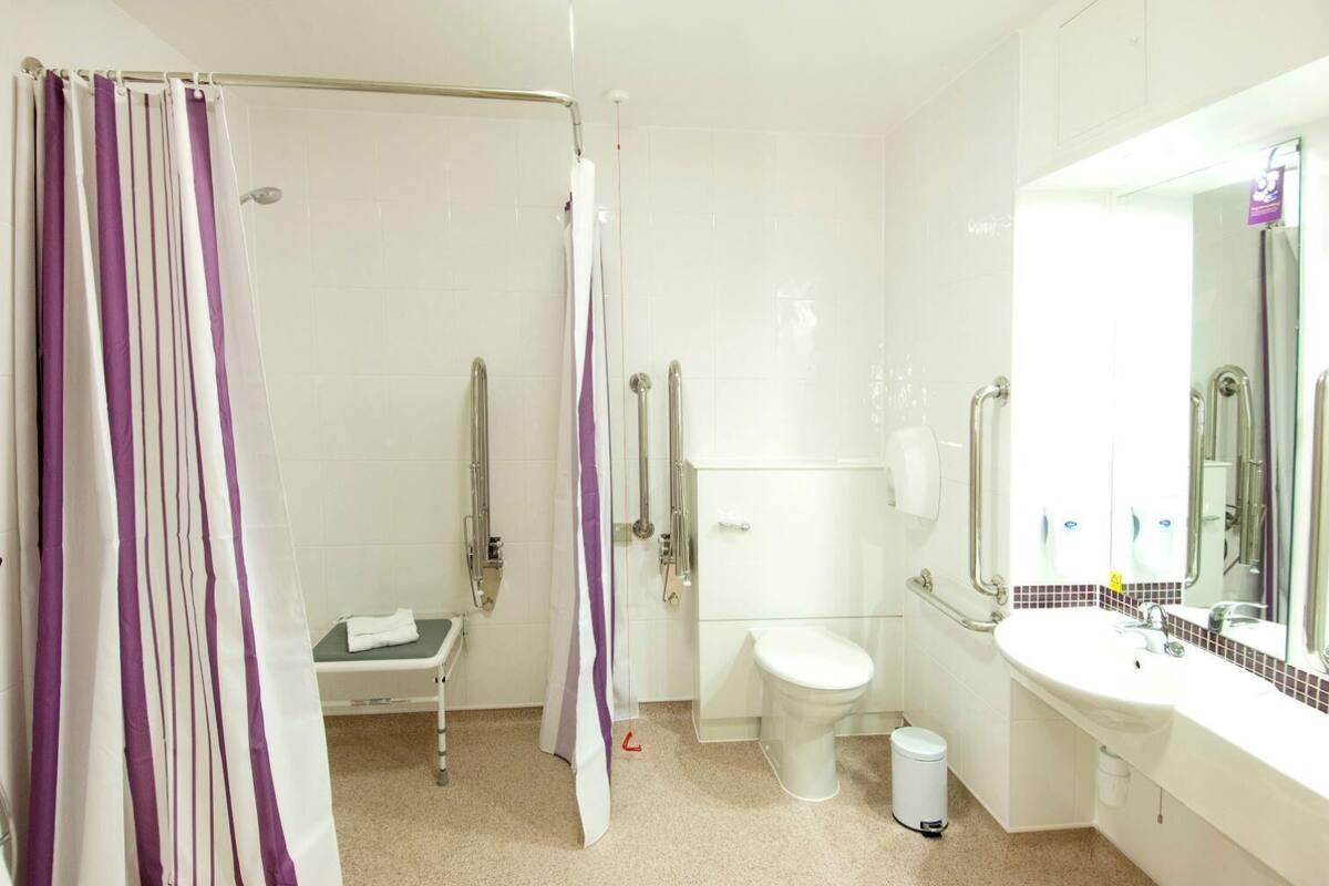 Un baño con ducha, inodoro y lavamanos, equipado con barras de apoyo y un banco accesible en silla de ruedas.