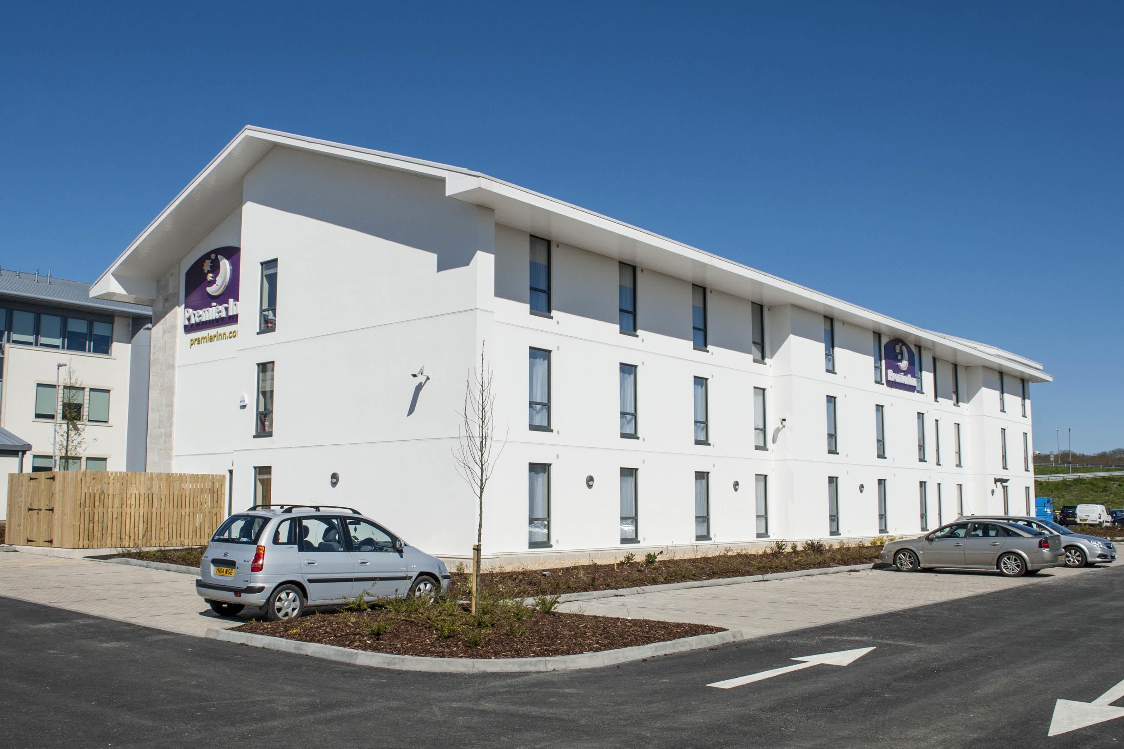 A modern building with a flat roof, multiple windows, and a sign indicating it is a Premier Inn hotel. There are cars parked in front.