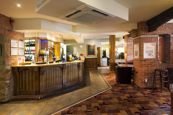 A bar area with a wooden counter, a brick wall, and a patterned carpet. - Premier Inn Bradford North - Bingley (Bingley)