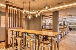 A modern restaurant with a long wooden table, bar stools, hanging pendant lights, and a view of the bar area. - Premier Inn Bristol Cribbs Causeway - M5 J17 (Bristol)
