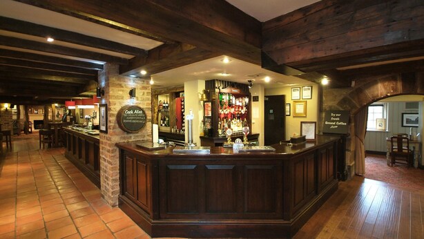 Zona de bar con vigas de madera, un pilar de piedra y una barra con diferentes botellas y vasos.
