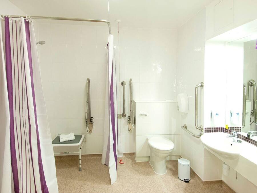 Ein Badezimmer mit Dusche, Toilette und Waschbecken, ausgestattet mit Haltegriffen und einer rollstuhlgerechten Sitzbank.