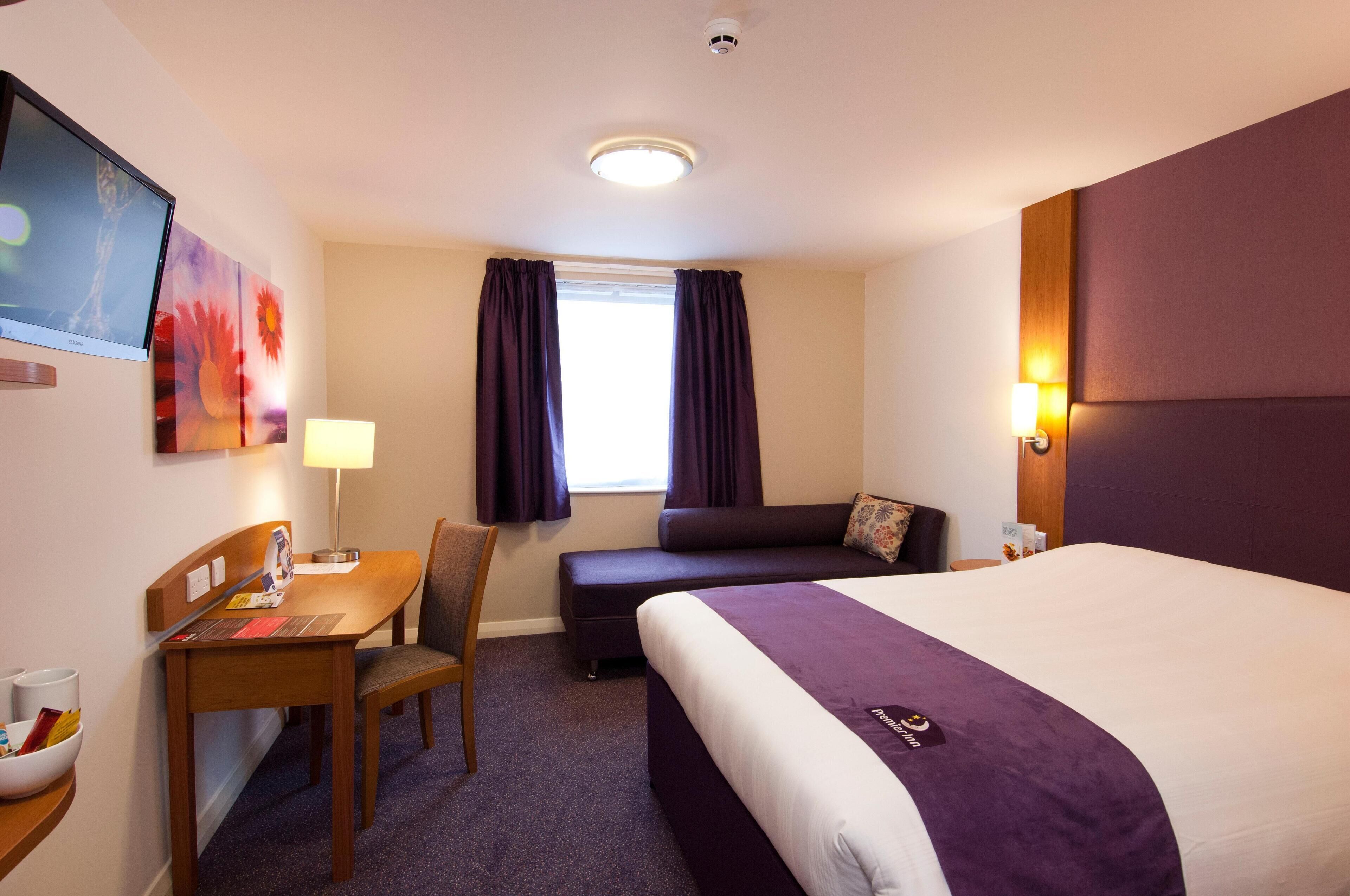 Quarto de hotel com cama, escrivaninha, cadeira, sofá, janela com cortinas e televisão presa na parede.